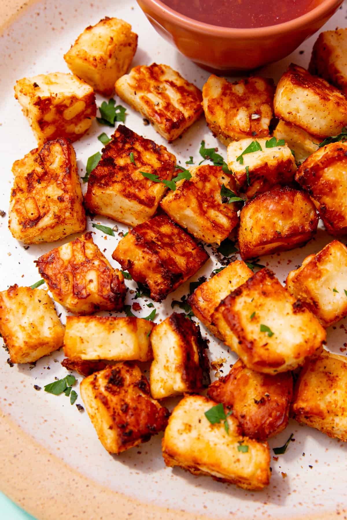 Golden browned halloumi cubes on a plate with some green herbs and a small bowl of chilli sauce.