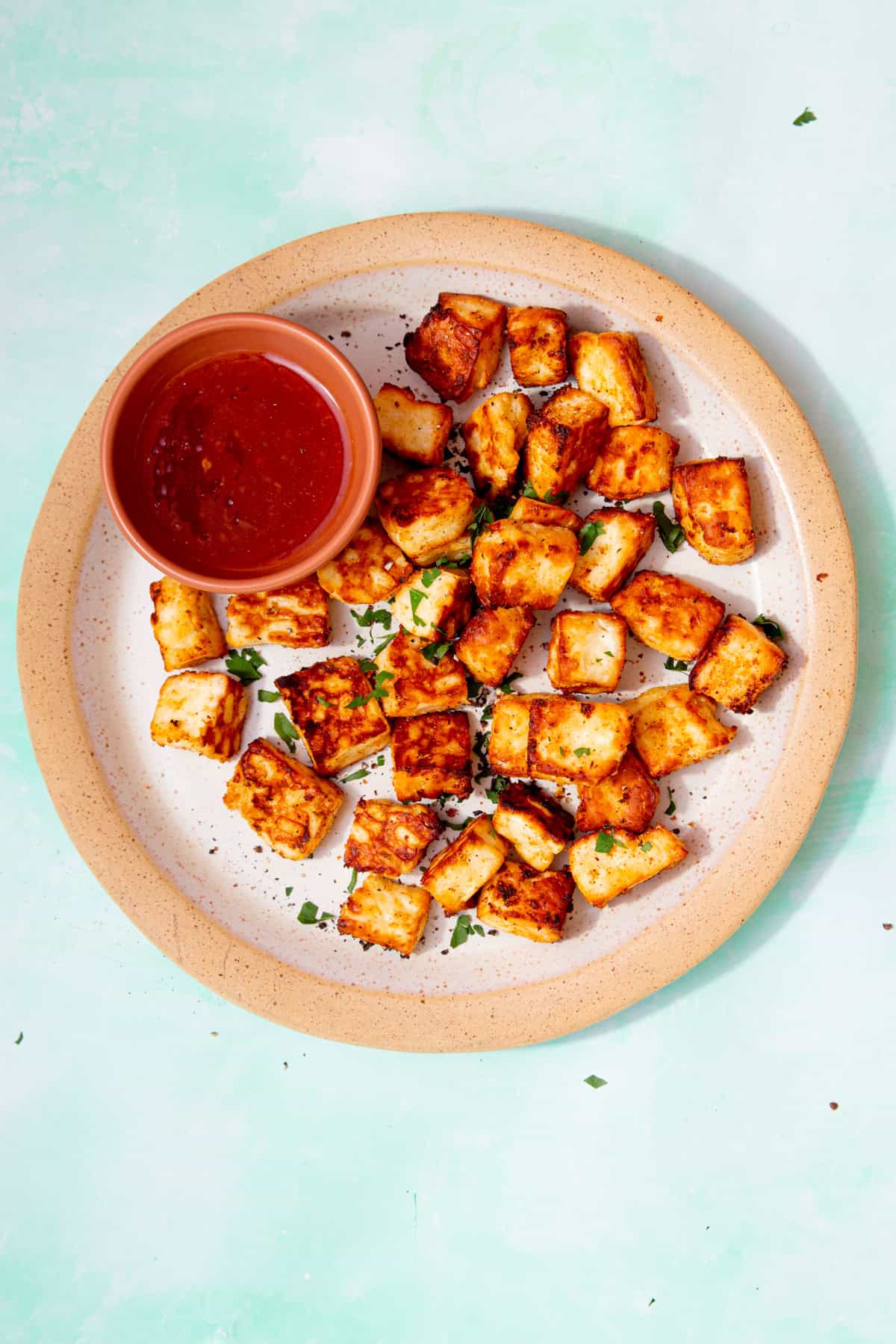 A plate filled with golden browned halloumi cubes with some green herbs and a small bowl of chilli sauce.