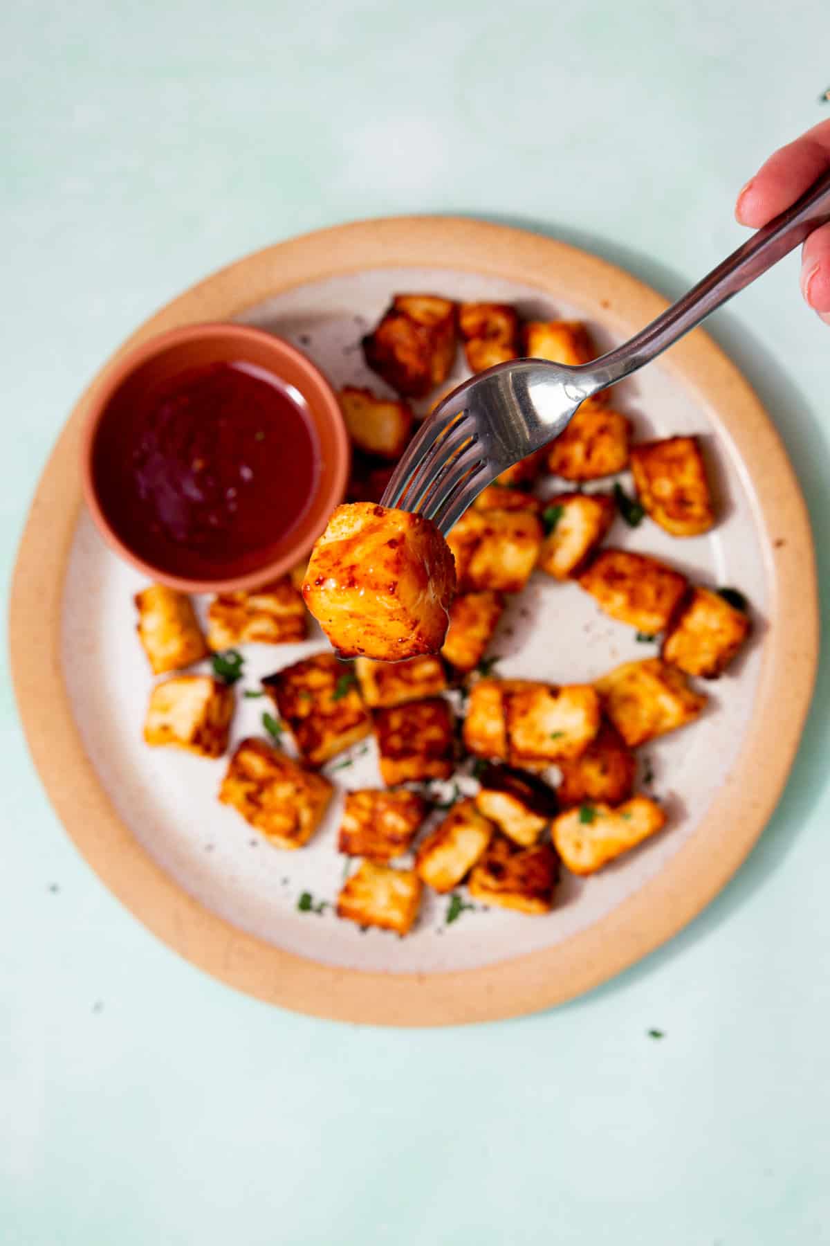 A plate filled with golden browned halloumi cubes with some green herbs and a small bowl of chilli sauce with a fork holding up a piece of halloumi.
