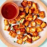 Golden browned halloumi cubes on a plate with some green herbs and a small bowl of chilli sauce on a pale blue background.