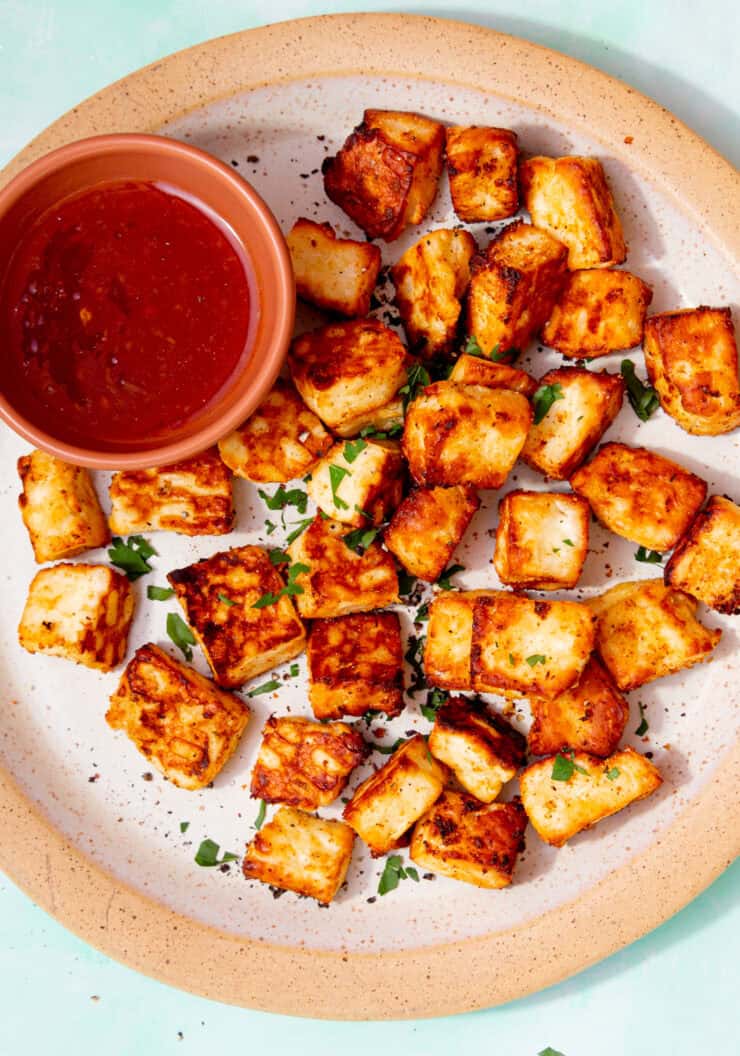Golden browned halloumi cubes on a plate with some green herbs and a small bowl of chilli sauce on a pale blue background.