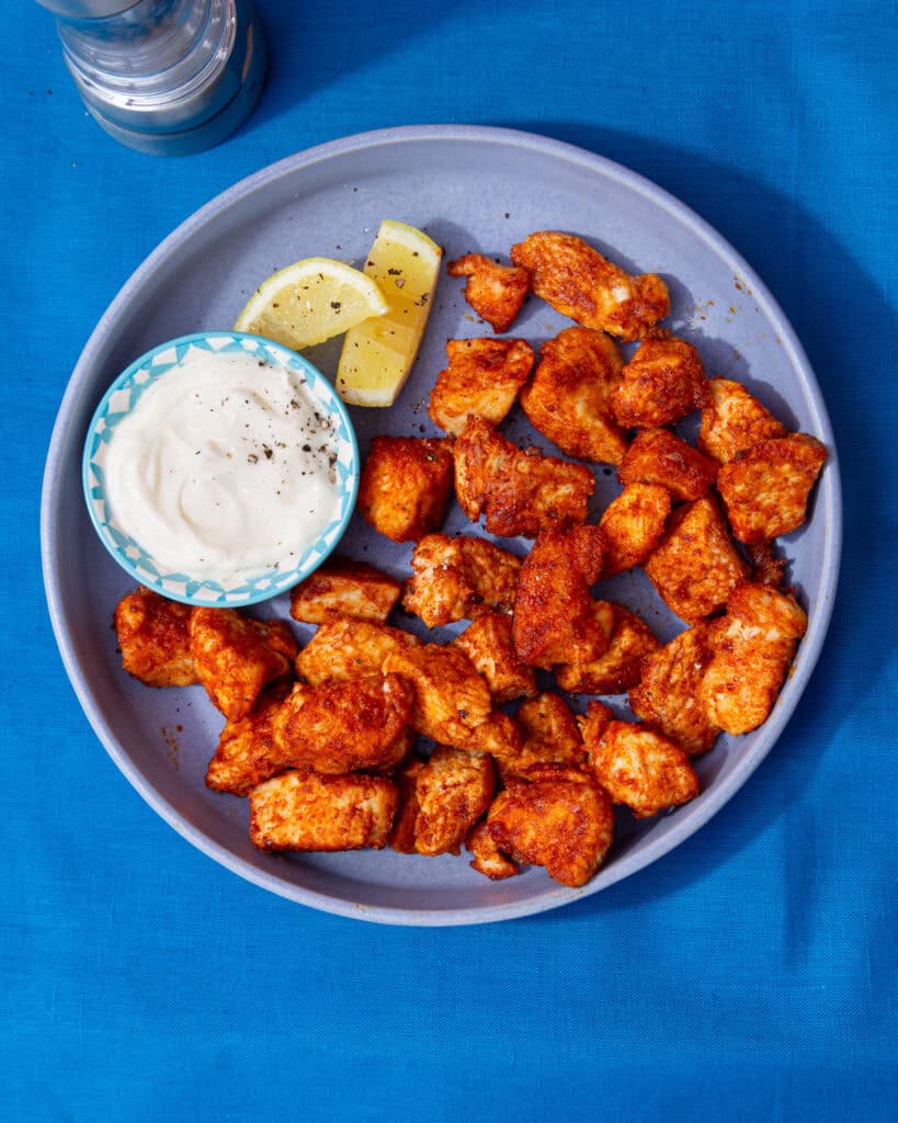 A blue bowl with golden browned and seasoned chicken ieces with wedges of lemon and a little bowl of mayonnaise.