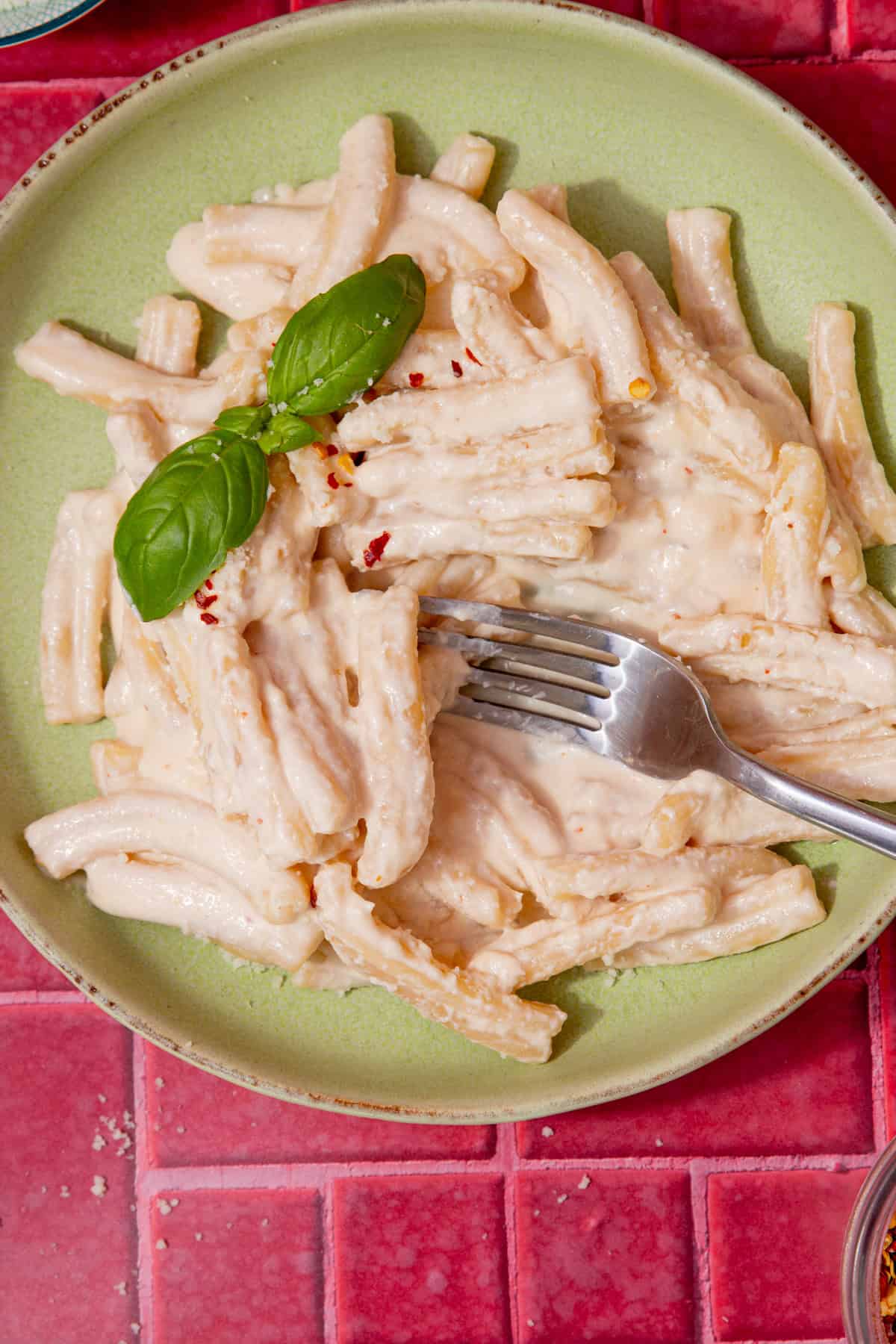A bowl with pasta and a creamy sauce with some fresh basil and a fork on a dark pink background.