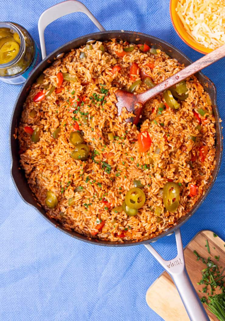 A large pan with a tomatoey coloured rice, with peppers and meat topped with jalapeños and mixed with a wooden spoon on a blue background.