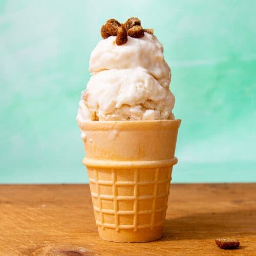 A cone on a wooden surface filled with white icecream with some brown toppings infront of a pale green background.