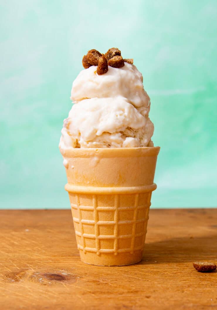 A cone on a wooden surface filled with white icecream with some brown toppings infront of a pale green background.