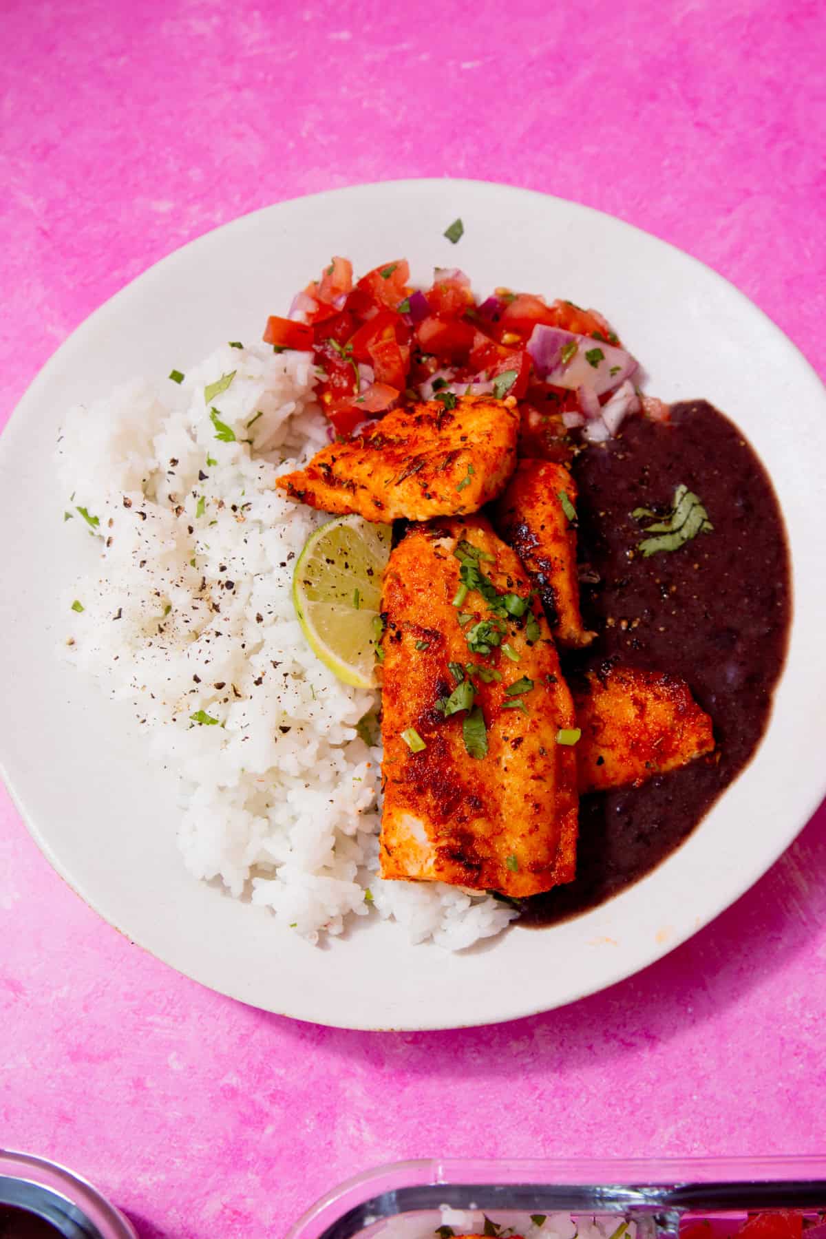 A plate with browned fish, black bean sauce, white rice and a tomato salad on a pink background.