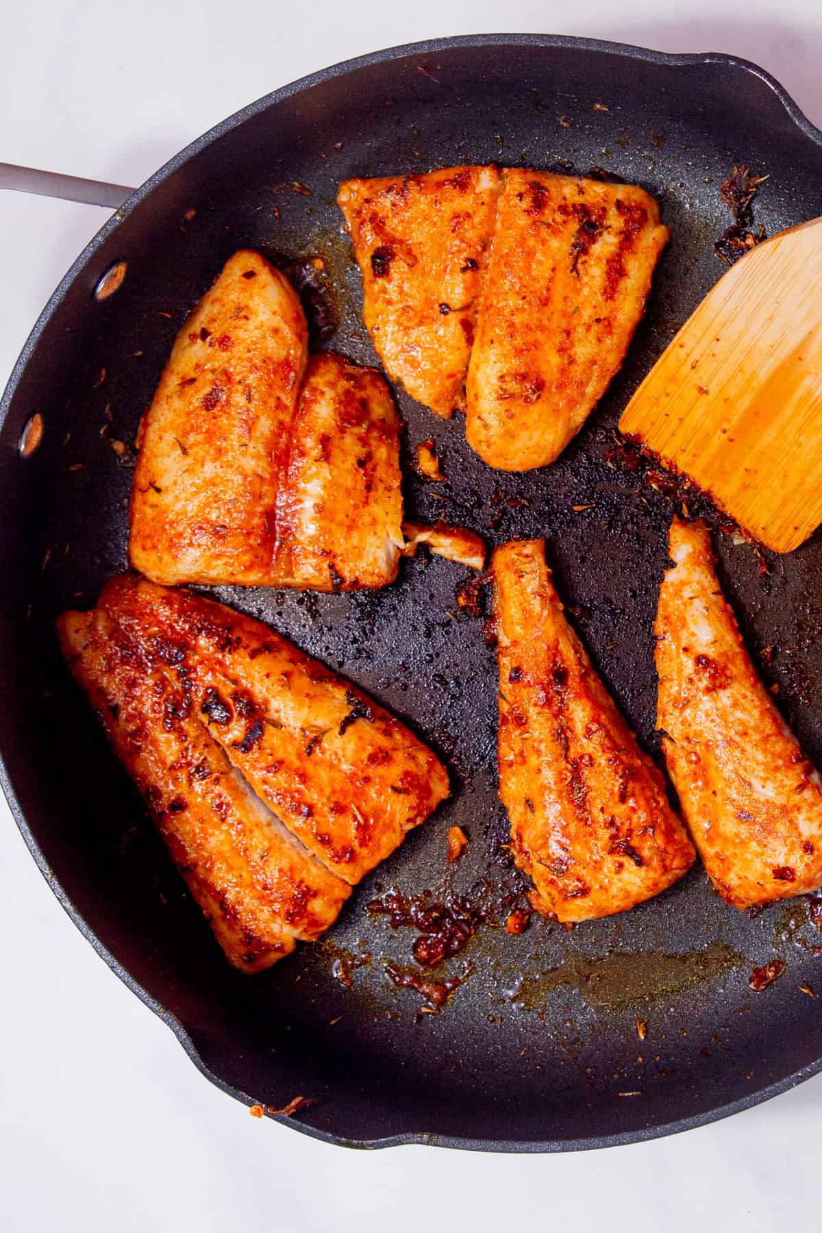 pieces of fish covered in a golden brown seasoning frying in a pan with a wooden spoon in partial view.