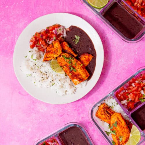 A plate with browned fish, black bean sauce, white rice and a tomato salad on a pink background with a few filled meal prep containers in view.