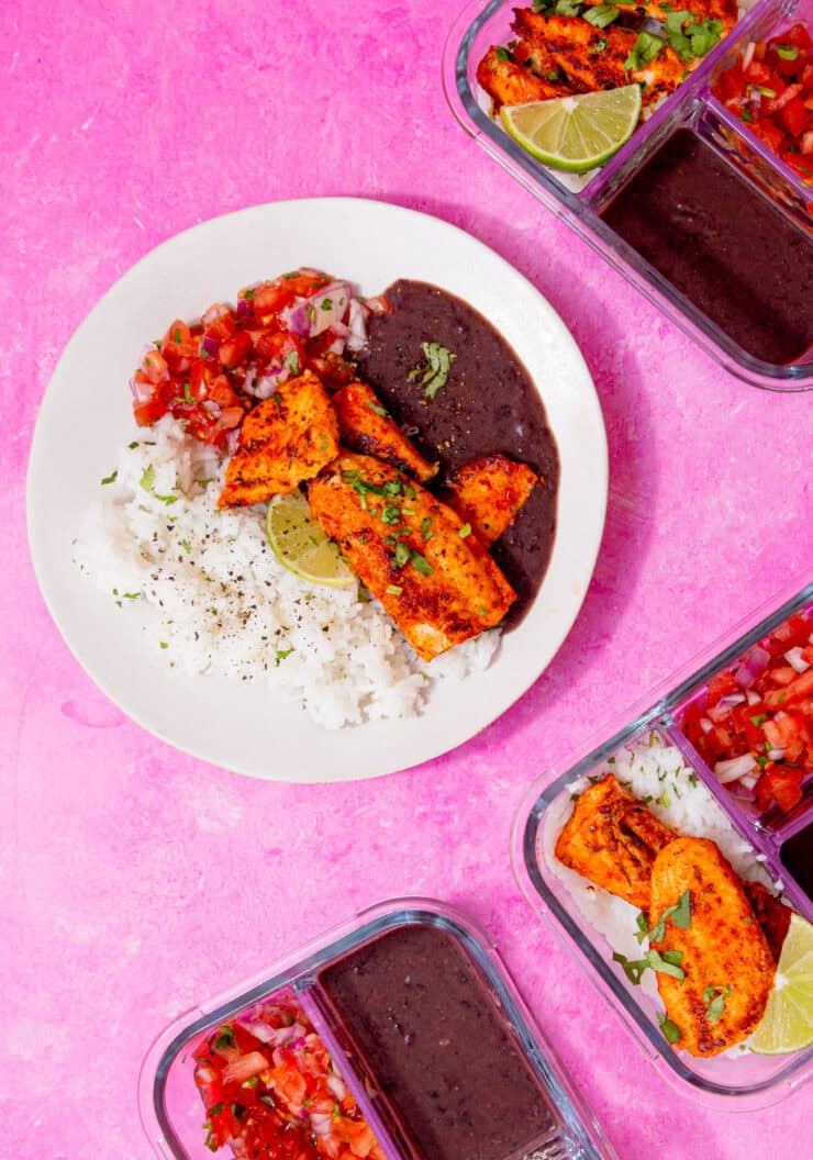 A plate with browned fish, black bean sauce, white rice and a tomato salad on a pink background with a few filled meal prep containers in view.