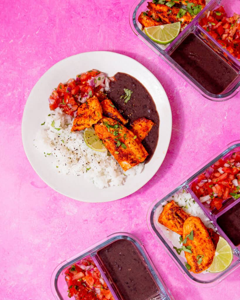 A plate with browned fish, black bean sauce, white rice and a tomato salad on a pink background with a few filled meal prep containers in view.