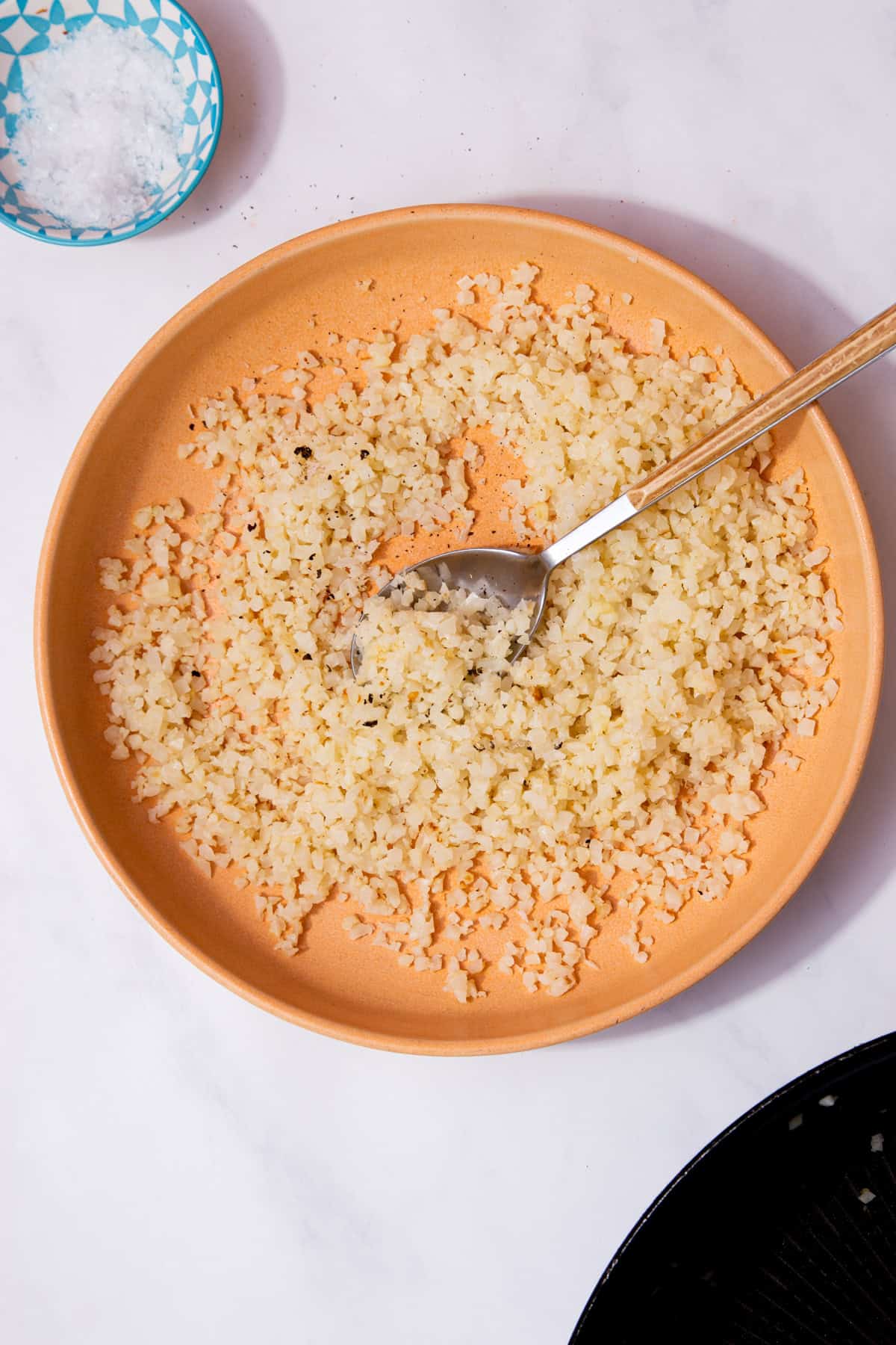 A pale tanned coloured bowl with cauliflower rice with a spoon on a white background next to a small bowl of salt.