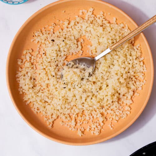 Caulflower rice in a pale tan bowl with a spoon on a white background next to a little bowl of salt.
