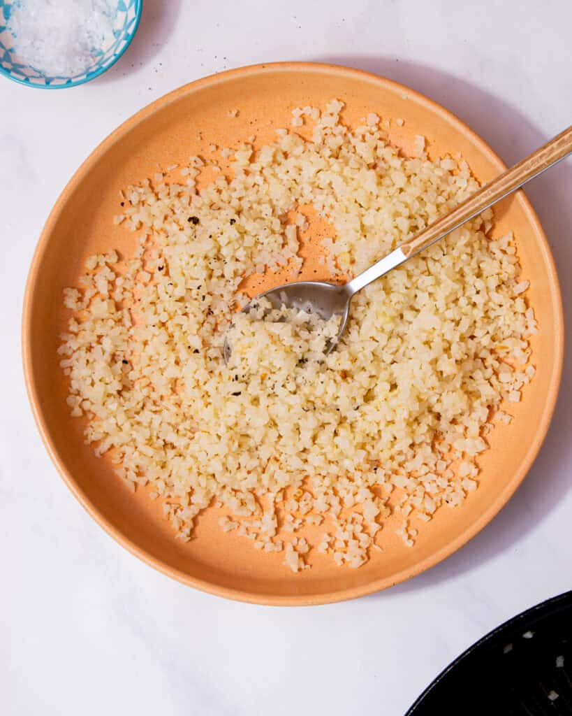 Caulflower rice in a pale tan bowl with a spoon on a white background next to a little bowl of salt.