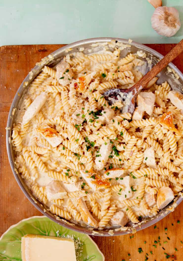 Fusilli cooked pasta in a pan with a creamy sauce, chicken pieces and some chopped chives with a wooden spoon on a wooden board next to a bowl with some parmesan.