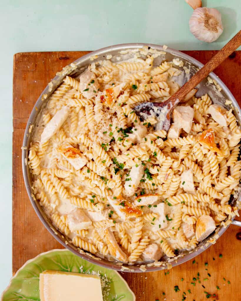 Fusilli cooked pasta in a pan with a creamy sauce, chicken pieces and some chopped chives with a wooden spoon on a wooden board next to a bowl with some parmesan.