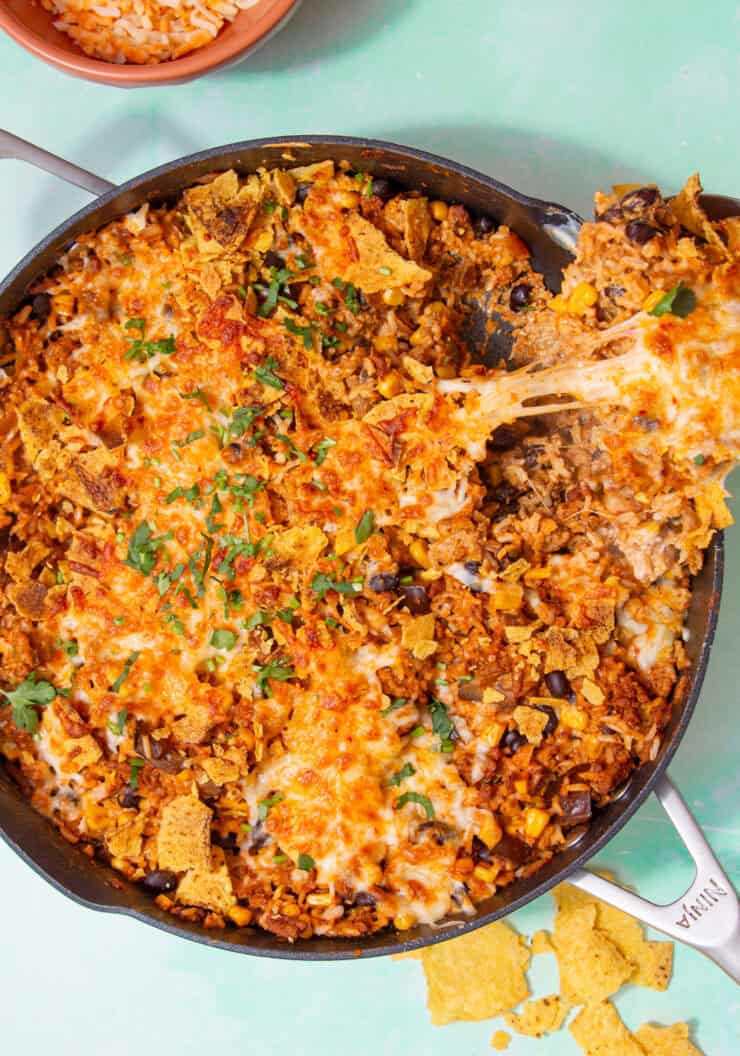 A large pan with golden brown cheesy topping and crumbled tortilla chips and some fresh herbs with aspoon taking out a portion of cheesy topping showing the rice, black beans and sweet corn and some tortilla chips lying next to the pan.