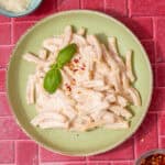 A pale green bowl with pasta and a creamy sauce with some fresh basil next to a small bowl of grated parmesan and a little bowl of chilli flakes on a dark pink background