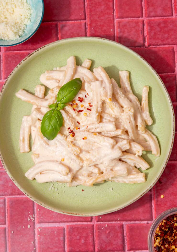 A pale green bowl with pasta and a creamy sauce with some fresh basil next to a small bowl of grated parmesan and a little bowl of chilli flakes on a dark pink background