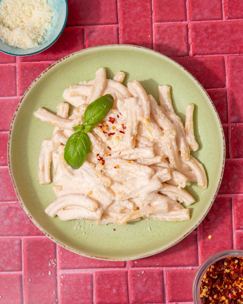 A pale green bowl with pasta and a creamy sauce with some fresh basil next to a small bowl of grated parmesan and a little bowl of chilli flakes on a dark pink background