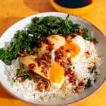 A bowl with eggs cut in quarters over rice with kale and roasted onions with chilli oil with a small bowl of soy sauce behind.
