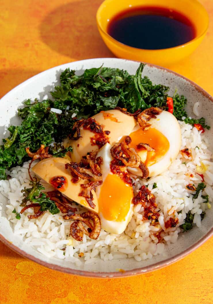 A bowl with eggs cut in quarters over rice with kale and roasted onions with chilli oil with a small bowl of soy sauce behind.