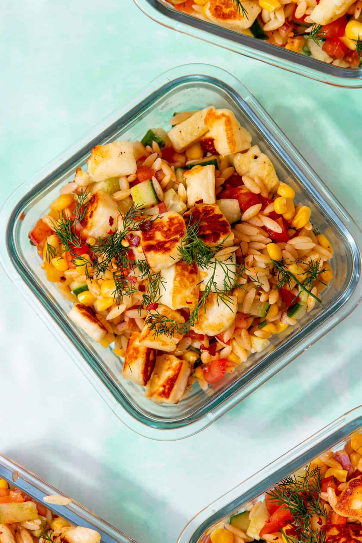 A square, glass meal prep container with orzo, salad and golden browned halloumi topped with some dill on a pale green background with other containers in partial view.