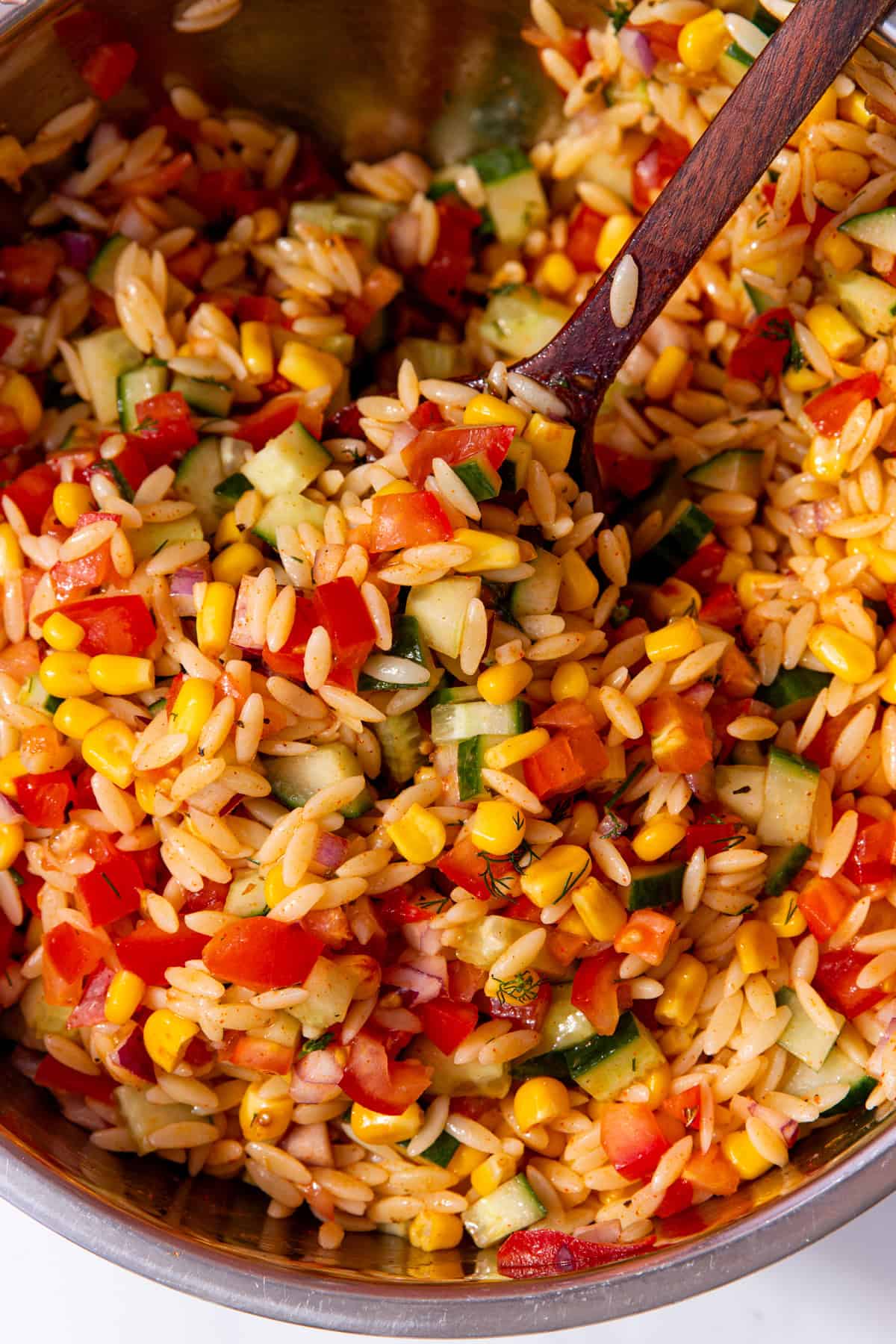 Orzo pasta salad with chopped tomatoes, cucumber, sweet corn and red onion in a metal bowl.
