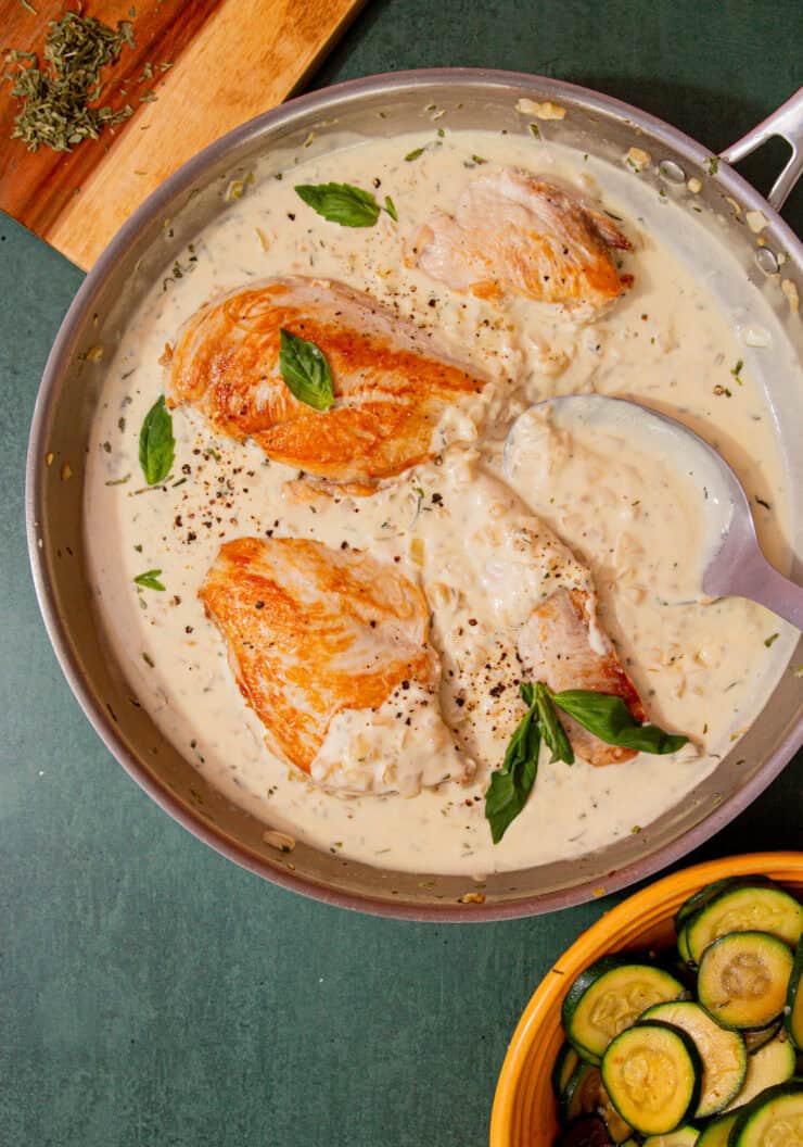 A large pan with golden browned chicken breast in a sauce with some fresh basil next to a bowl of courgette rouds and some herbs on a chopping board.