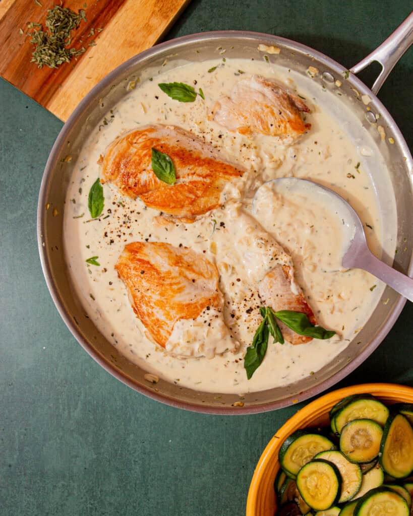 A large pan with golden browned chicken breast in a sauce with some fresh basil next to a bowl of courgette rouds and some herbs on a chopping board.