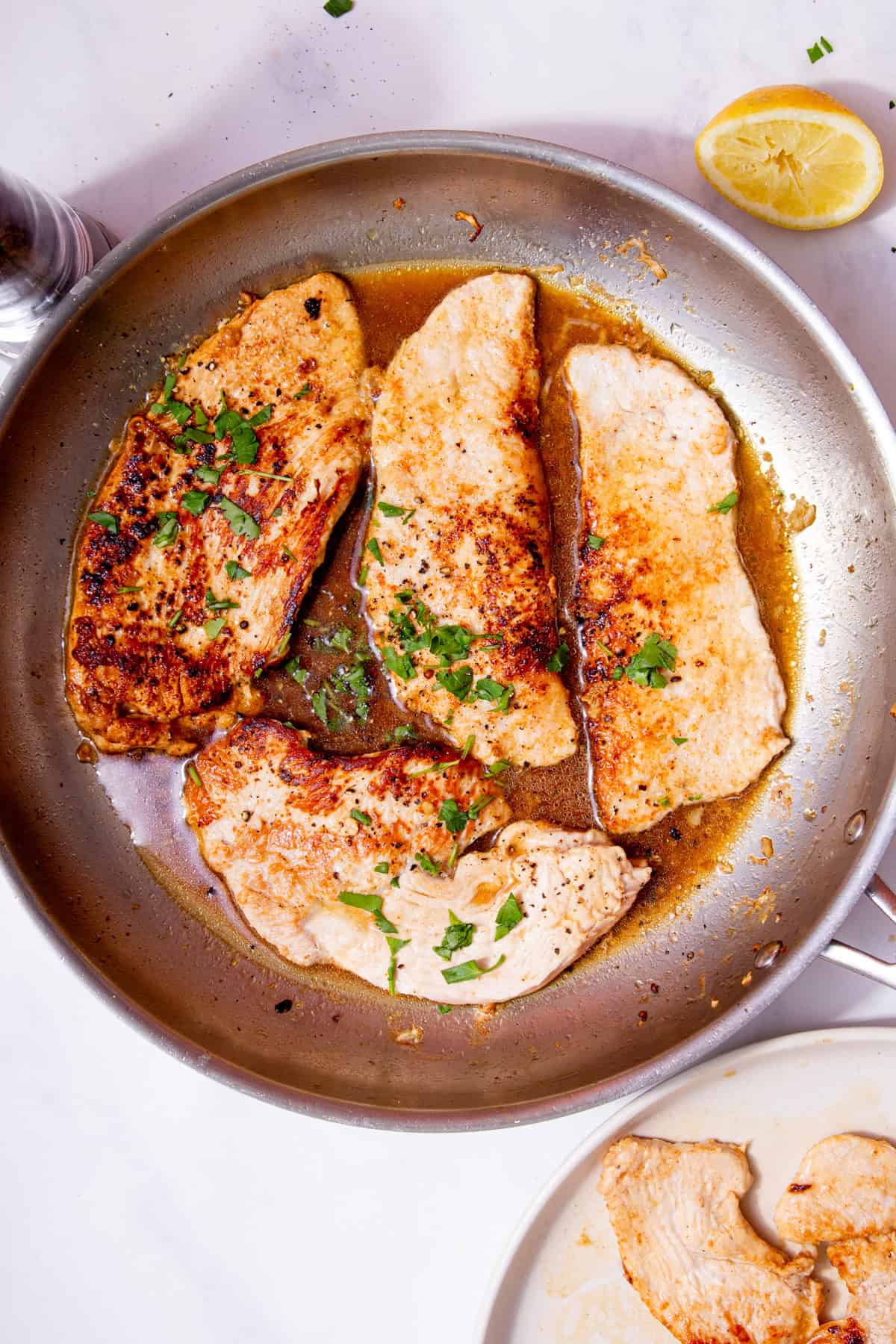 Turkey steaks in a sauce in a stainless steel frying pan and fresh green herbs with a half a used lemon on a white background.