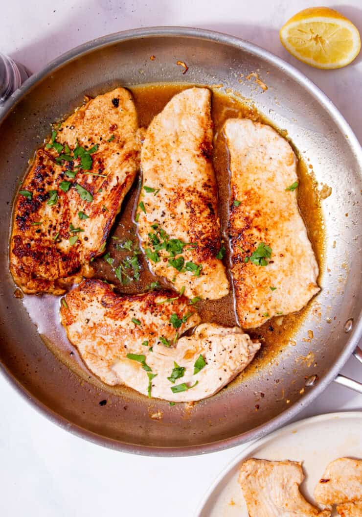 Turkey steaks browned in a large stainless steel frying pan with some brown stock, fresh herbs and a lemon wedge.