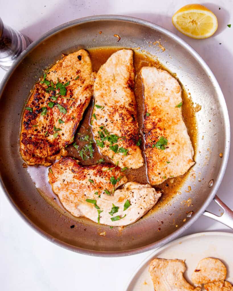 Turkey steaks browned in a large stainless steel frying pan with some brown stock, fresh herbs and a lemon wedge.