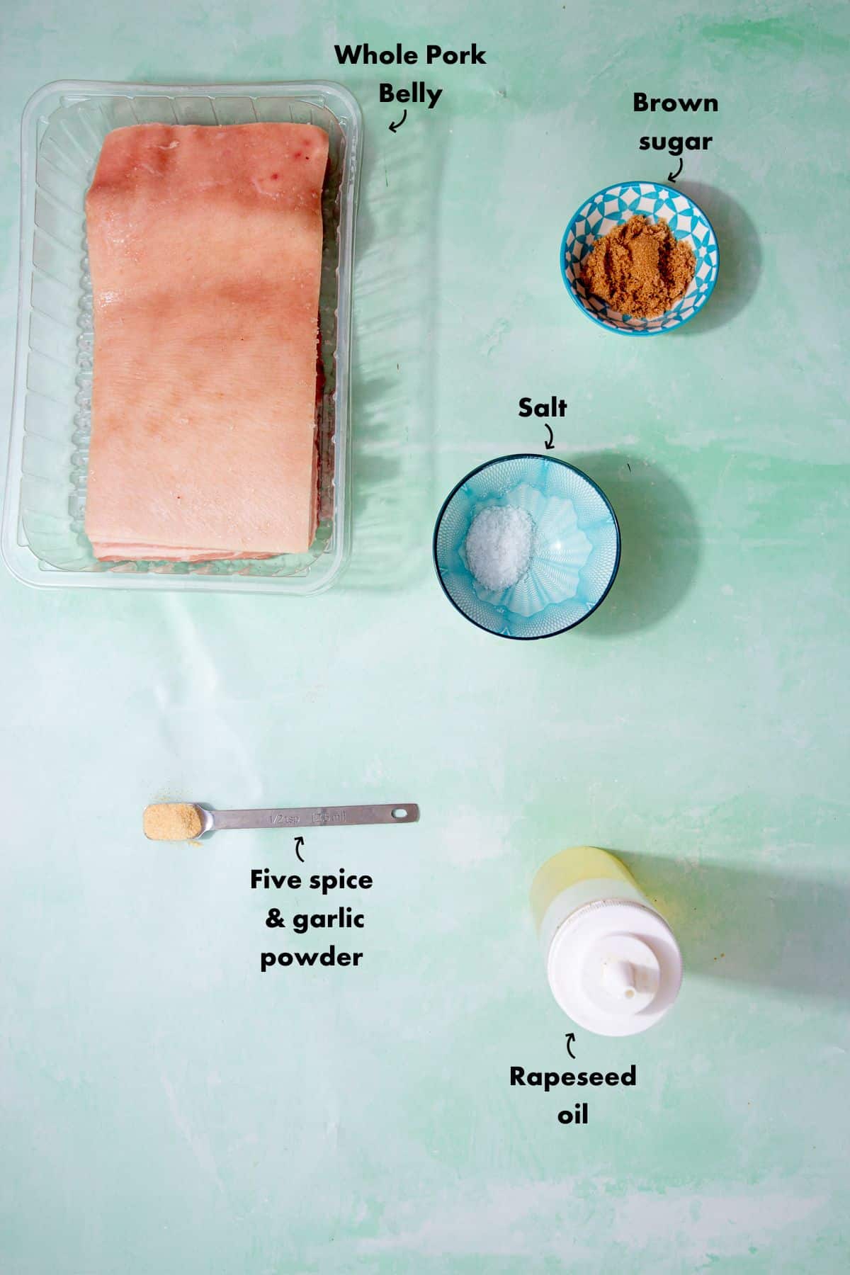 Ingredients to make a whole pork belly recipe with brown sugar, spcies and salt laid out on a pale blue background and labelled.