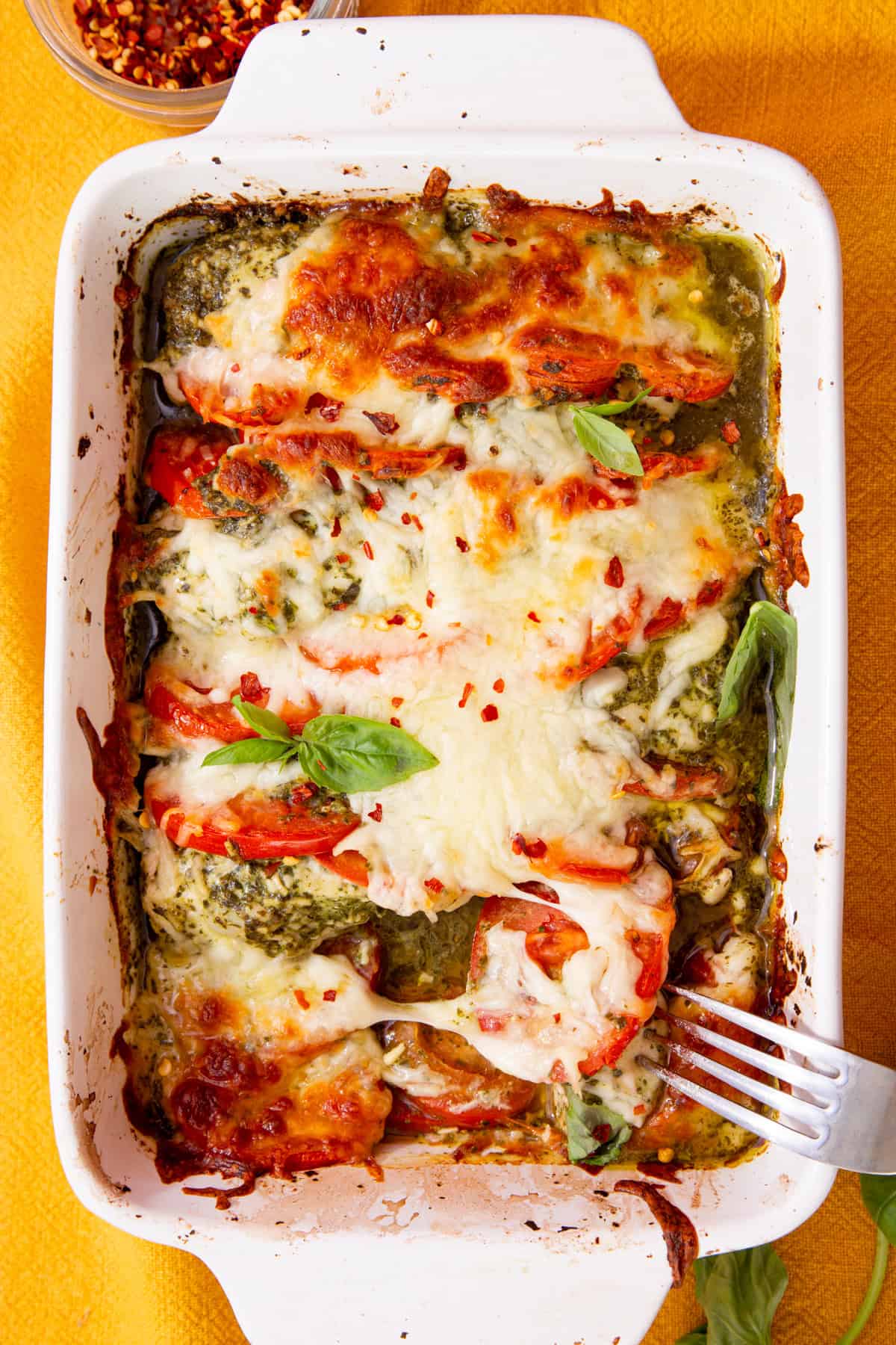 A white rectangular baking dish filled with baked, browned pesto chicken covered in golden browned mozzarella cheese with some fresh basil.