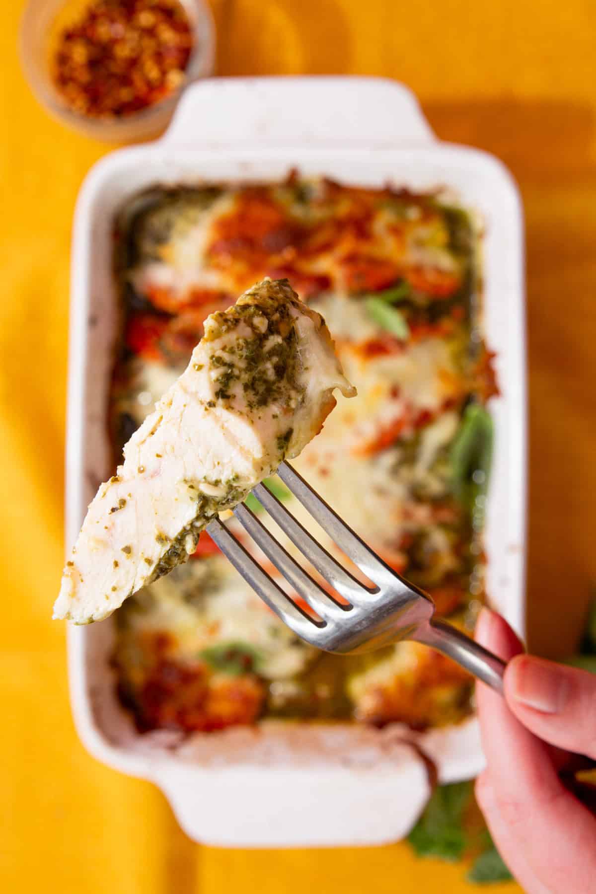 A white rectangular baking dish filled with baked, browned pesto chicken covered in golden browned mozzarella cheese with some fresh basil with a piece of chicken being held up with a fork.