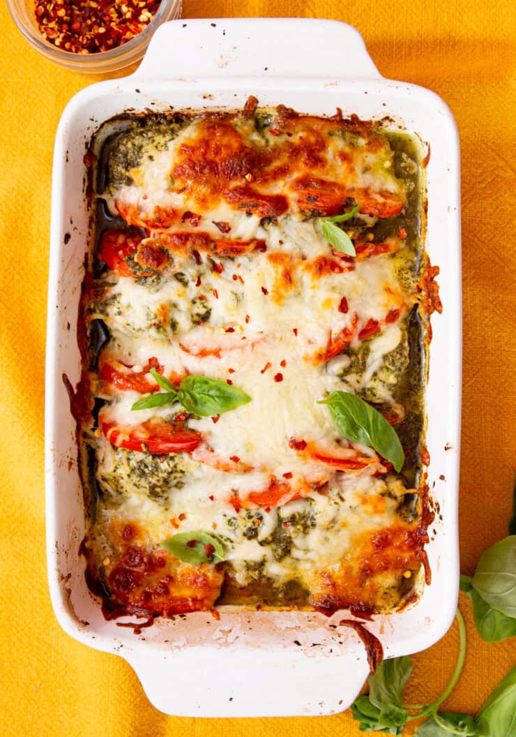 A white, rectangular baking dish with chicken and tomatoes covered in melted golden browned mozzarella with some fresh basil.