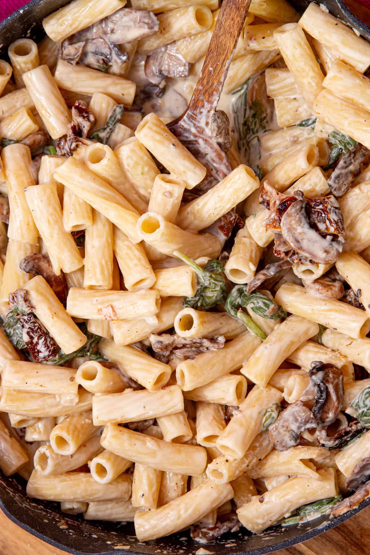 Rigatoni pasta with a creamy mushroom sauce with wilted spinach and a wooden spoon.