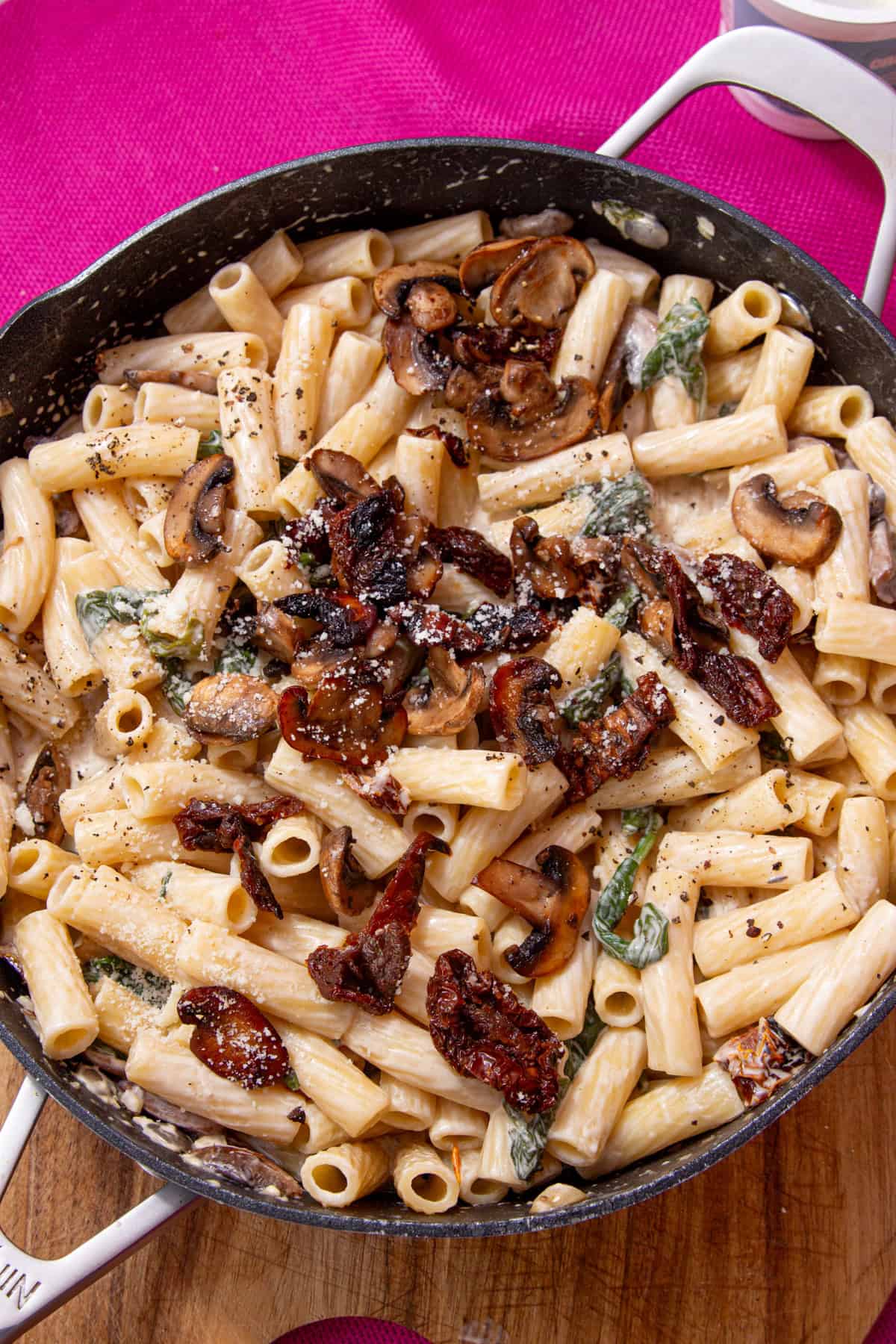 Rigatoni pasta with a creamy mushroom sauce with wilted spinach and a wooden spoon in a large pan on a pink background.