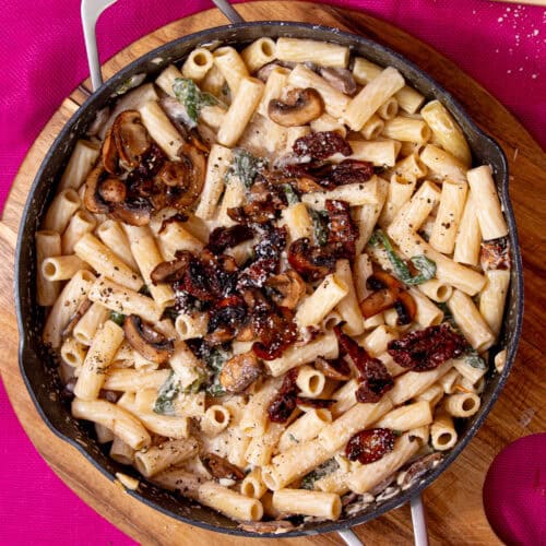 A large pan with a creamy mushroom pasta topped with mushrooms and sun-dried tomatoes on a pink back ground.