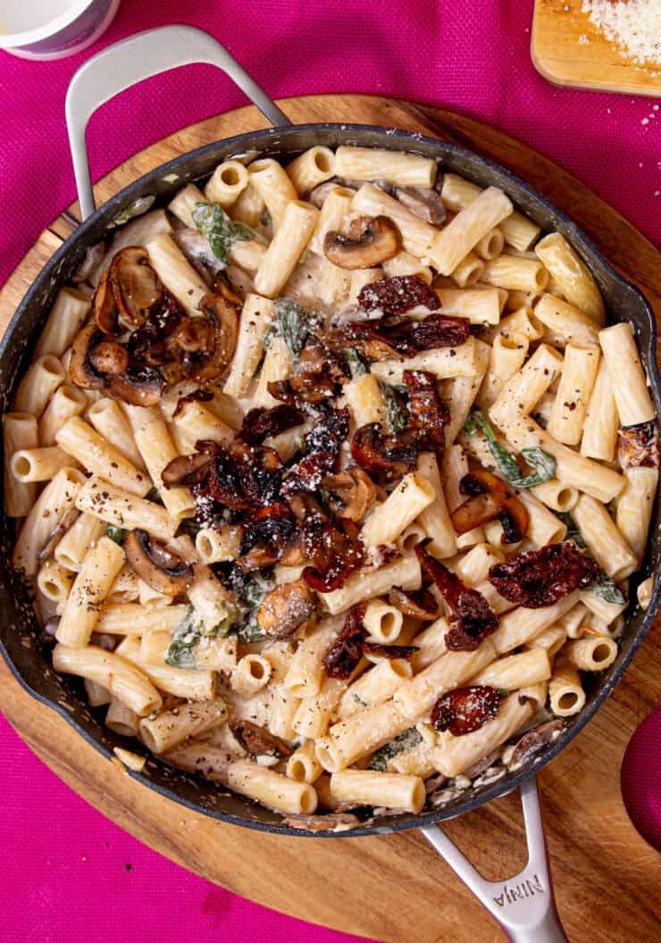 A large pan with a creamy mushroom pasta topped with mushrooms and sun-dried tomatoes on a pink back ground.