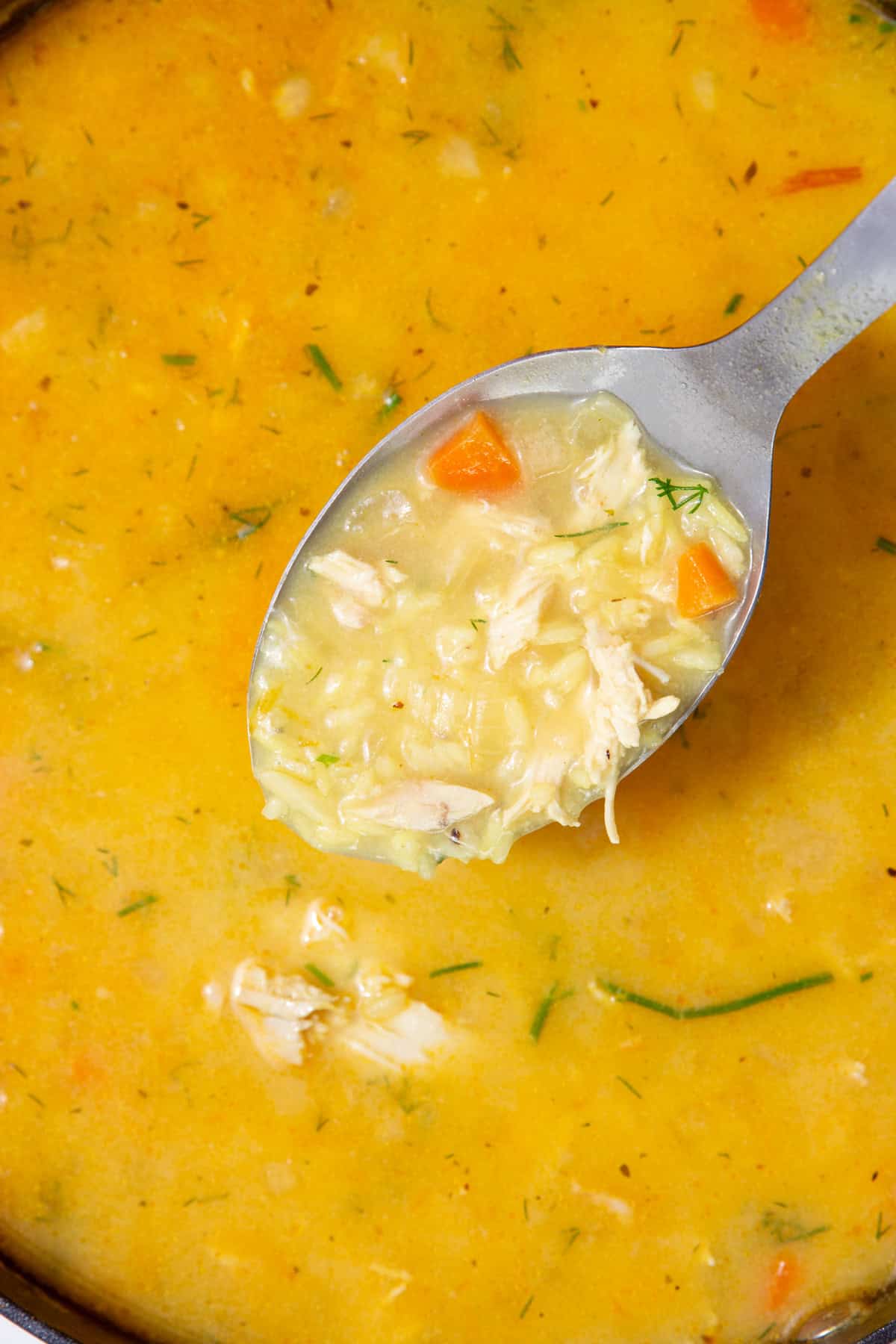 A yellowish coloured soup with a stainless steel spoonwith rice, carrot and onions.