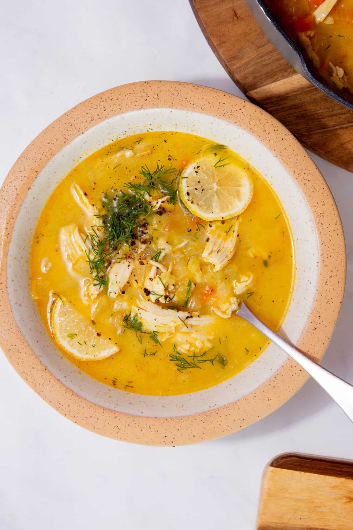 A bowl with a yellowish coloured soup with chicken, topped with slices of lemon  and fresh dill on a white background.