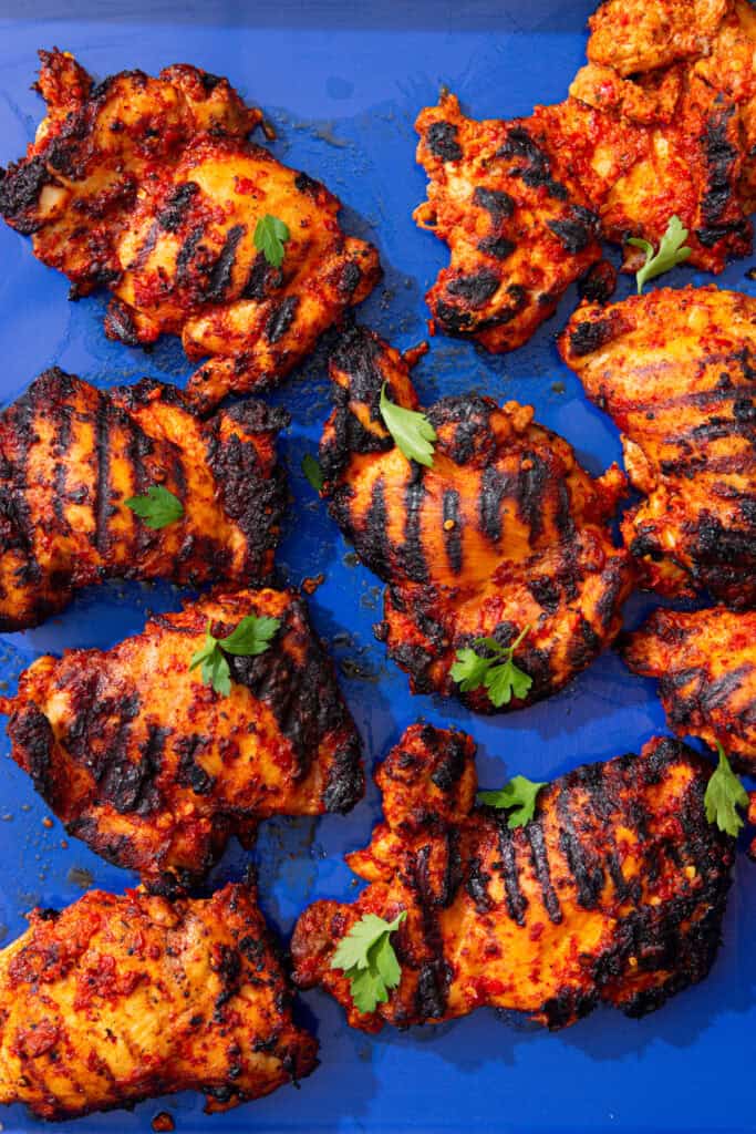 Browned and charred chicken thighs laid out on a blue tray with some green herbs.