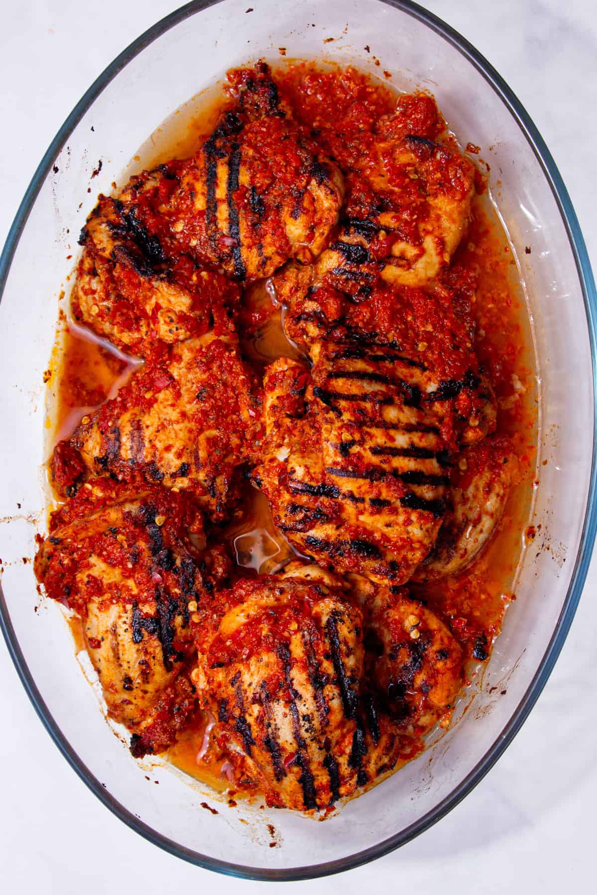 Charred chicken thighs in a marinade cooked in a glass oval dish on a white background.