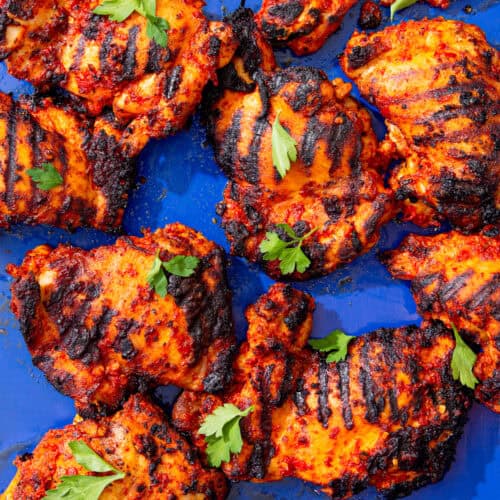 Lots of marinated and charred chicken thighs on a blue tray with some fresh parsley.