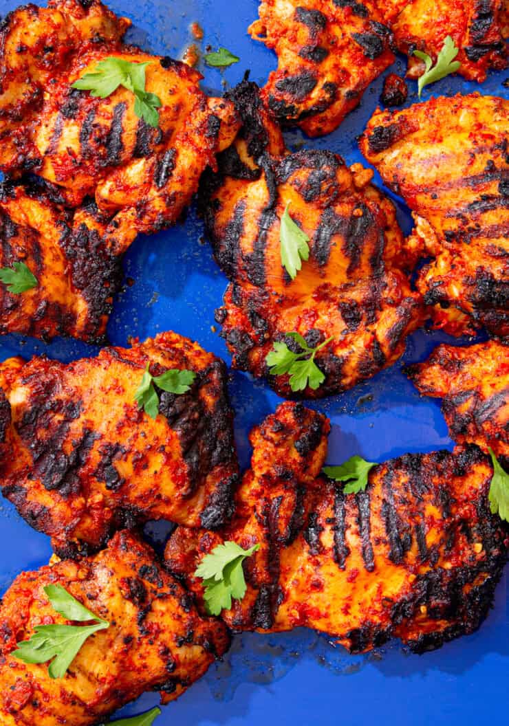 Lots of marinated and charred chicken thighs on a blue tray with some fresh parsley.