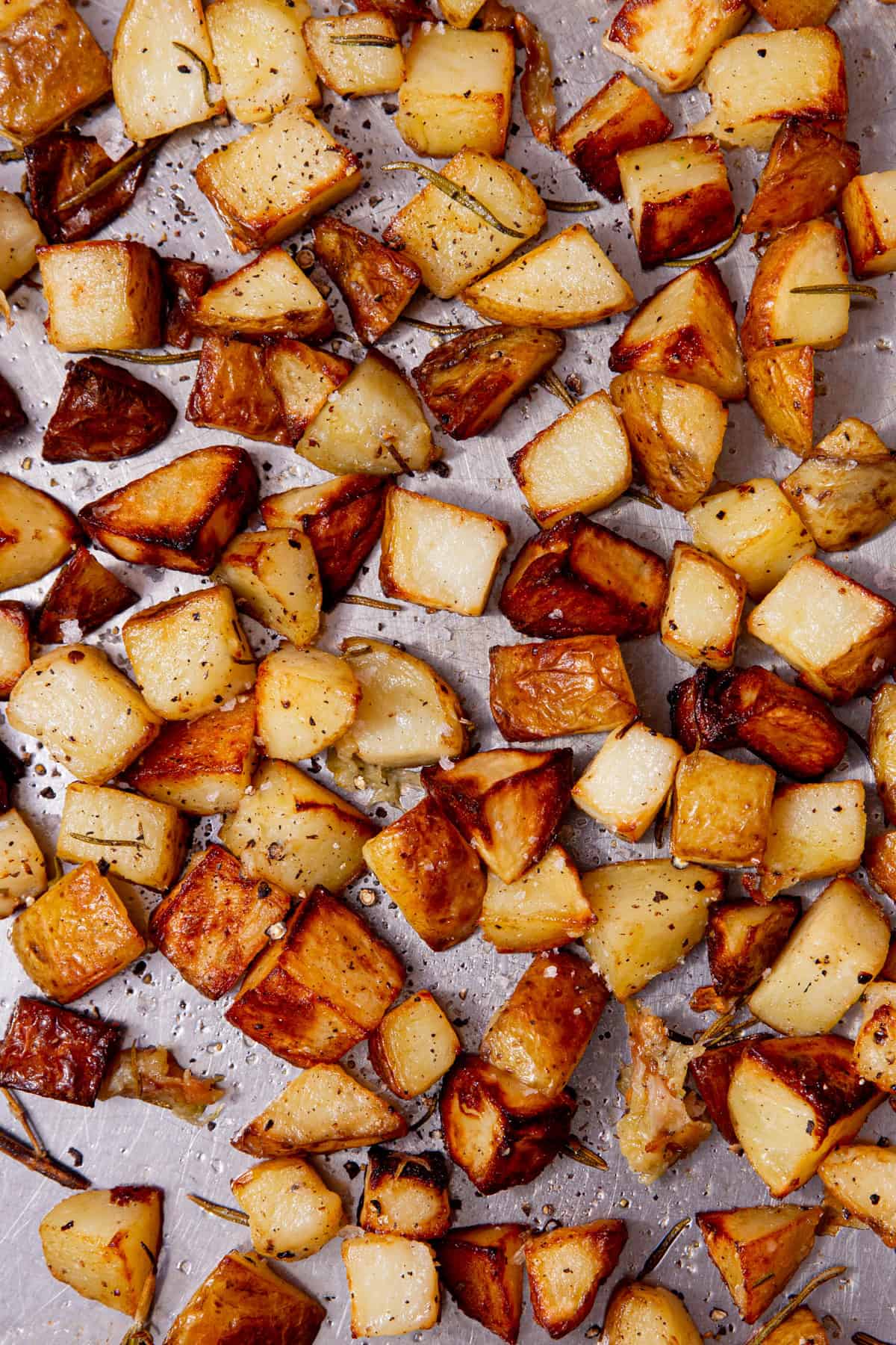 A large stainless steel baking tray filled with with browned, roasted potato cubes with some seasoing and rosemary sprigs.