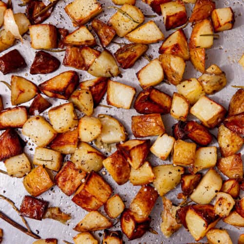 A large stainless steel baking tray with browned, roasted potato cubes with some seasoning and rosemary sprigs.