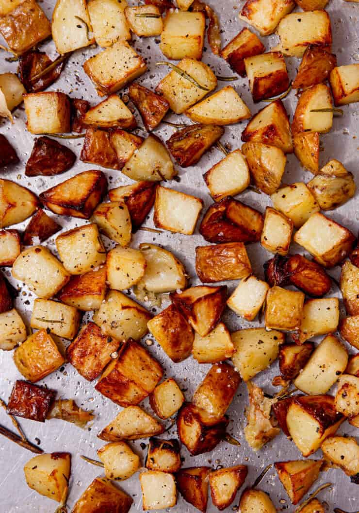 A large stainless steel baking tray with browned, roasted potato cubes with some seasoning and rosemary sprigs.